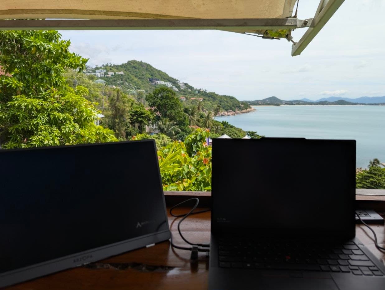 A laptop and external monitor set up on a balcony desk overlooking a hillside and the sea
